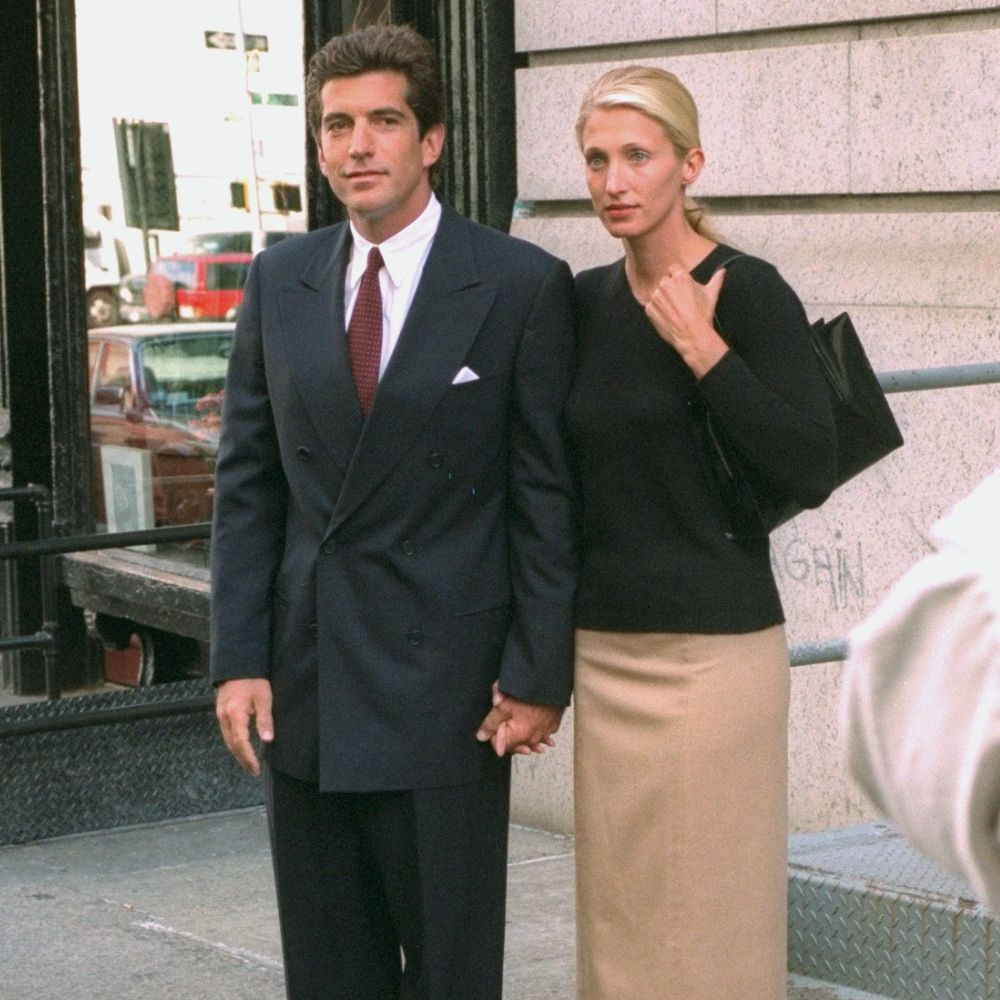 Carolyn Bessette-Kennedy wears a knee-length pencil skirt with brown knee-high boots and a black v-neck knit.
