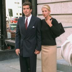 Carolyn Bessette-Kennedy wears a knee-length pencil skirt with brown knee-high boots and a black v-neck knit.