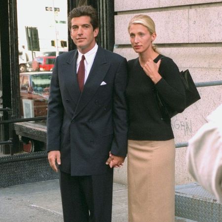 Carolyn Bessette-Kennedy wears a knee-length pencil skirt with brown knee-high boots and a black v-neck knit.