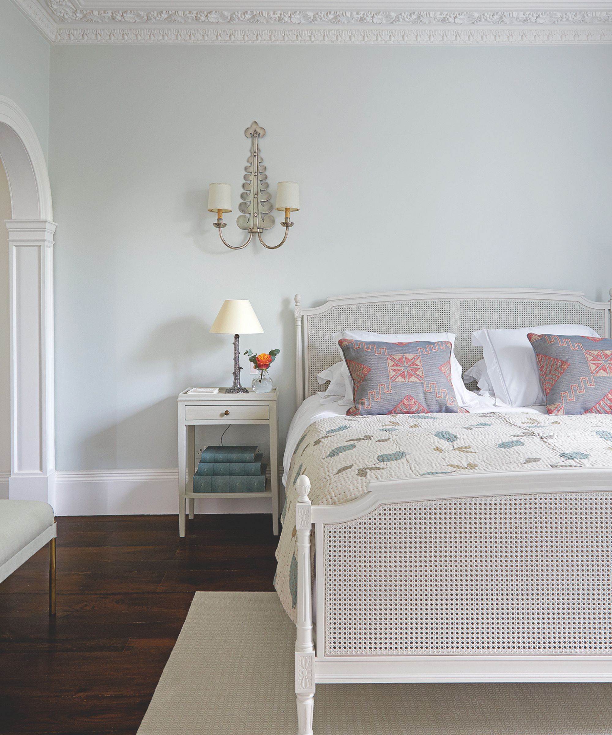 A blue bedroom with white cornicing and baseboards, dark wooden flooring, a white rug, white rattan bed, and white bedside table.