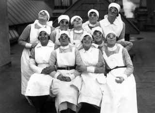 A group of ten nurses wearing their uniform and round dark glasses all smile and look up to the sky.