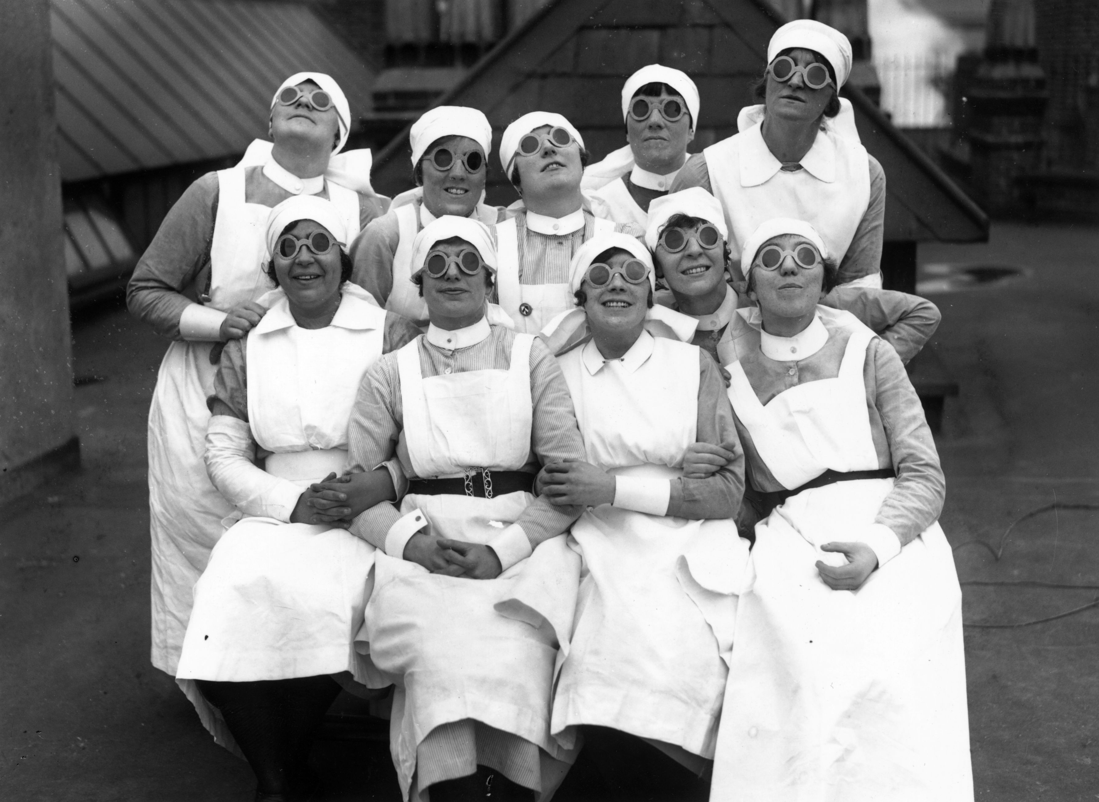 A group of ten nurses wearing their uniform and round dark glasses all smile and look up to the sky.