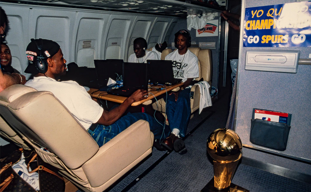 San Antonio Spurs players Tim Duncan, David Robinson, Malik Rose, and Sean Elliott sit at a table on an airplane, with laptops connected by ethernet cables. Their championship trophy is visible in the foreground.