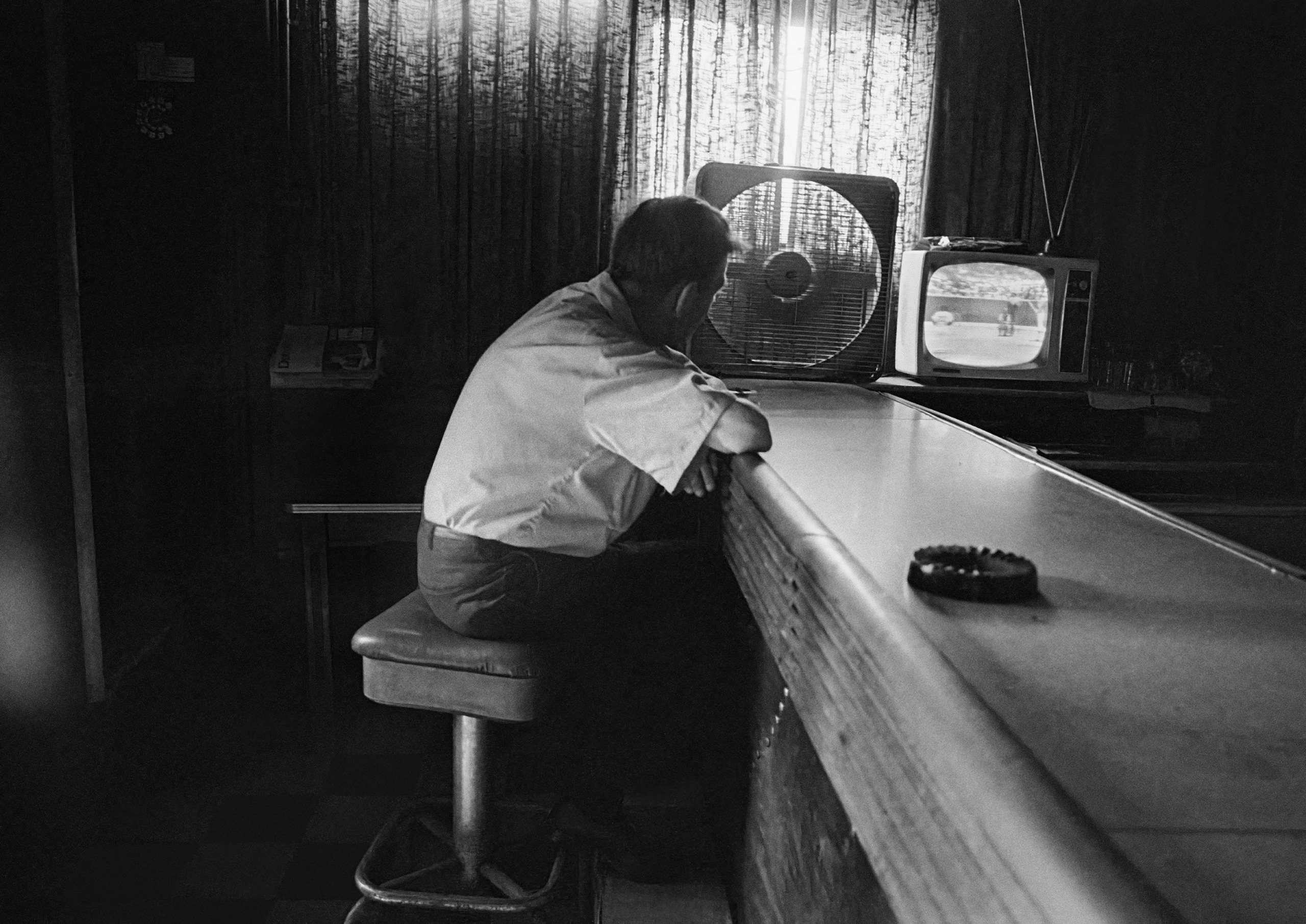 A man sits alone on a stool at a dimly lit bar, leaning on the counter beside a box fan as he watches a baseball game on a small portable television.
