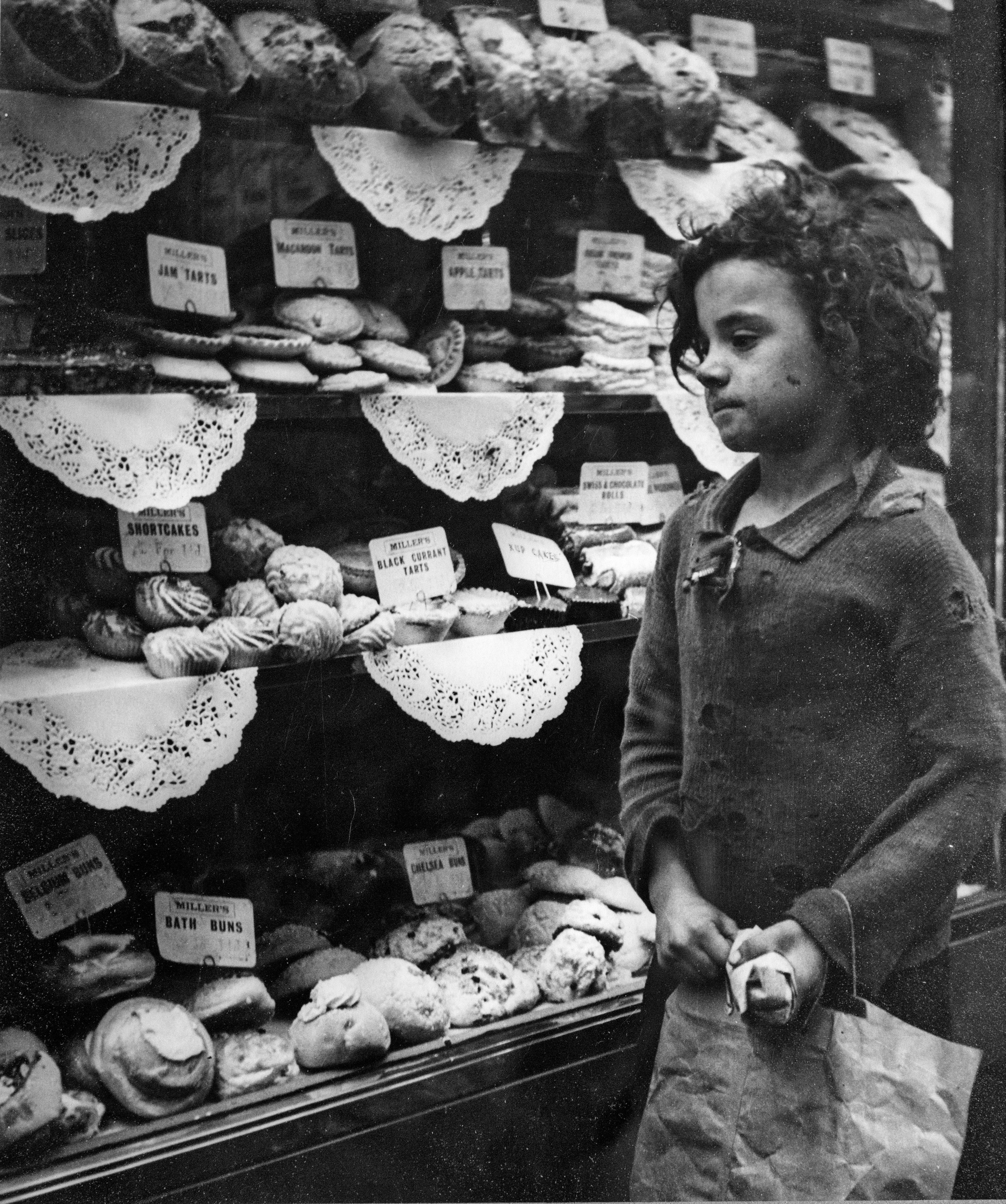 A young child with curly hair and worn, patched clothing stands before a bakery window crammed with pastries and cakes on lace-doily-lined shelves, including jam tarts, shortcakes and Chelsea buns. The child holds a paper bag and gazes at the display with a quietly absorbed expression.