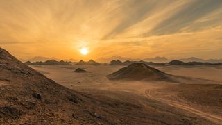 A photo of a sunset in the desert. It is a flat sandy lanscape, with several small hills popping up here and there. You can just make out two people who have climbed up a mountain in the distance. The location is Qesm Safaga, Red Sea Governorate, Egypt.