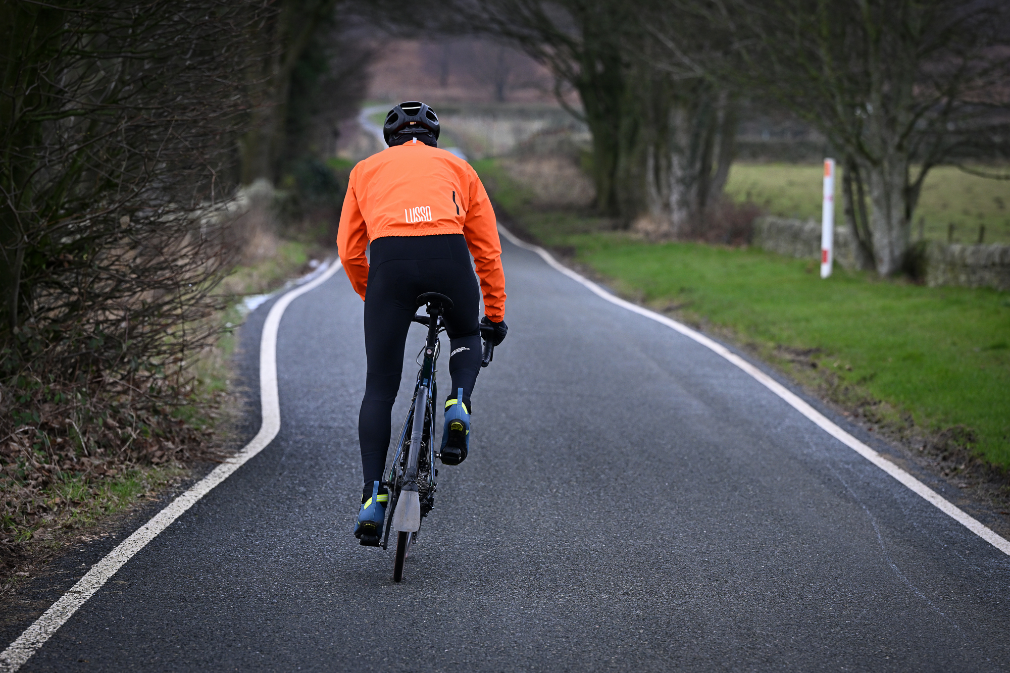 Man wearing black tights, blue shoes and an orange jacket riding a green road bike away from the camera up a country lane
