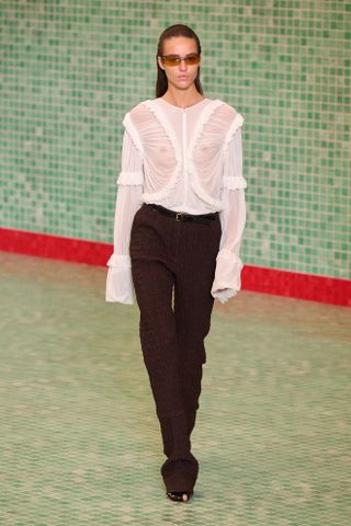 A model at the spring/summer 2025 Tory Burch show wearing a sheer white draped blouse with brown trousers, sunglasses, and peep-toe heels.