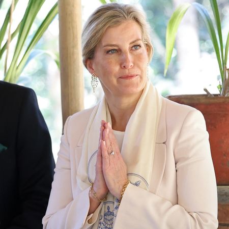 Duchess Sophie wearing a cream blazer and scarf holding her hands in prayer and looking to the right smiling with palm leaves behind her