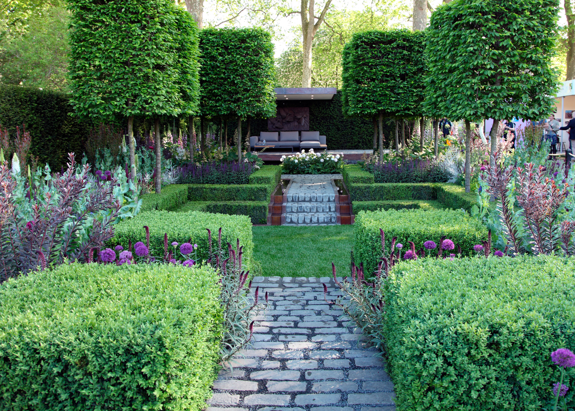 A garden with a brick path, box hedges, and four pleached trees