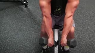 Trainer from Anabolic Aliens sitting down holding two dumbbells with arms extended and tapping ends of the dumbbells together