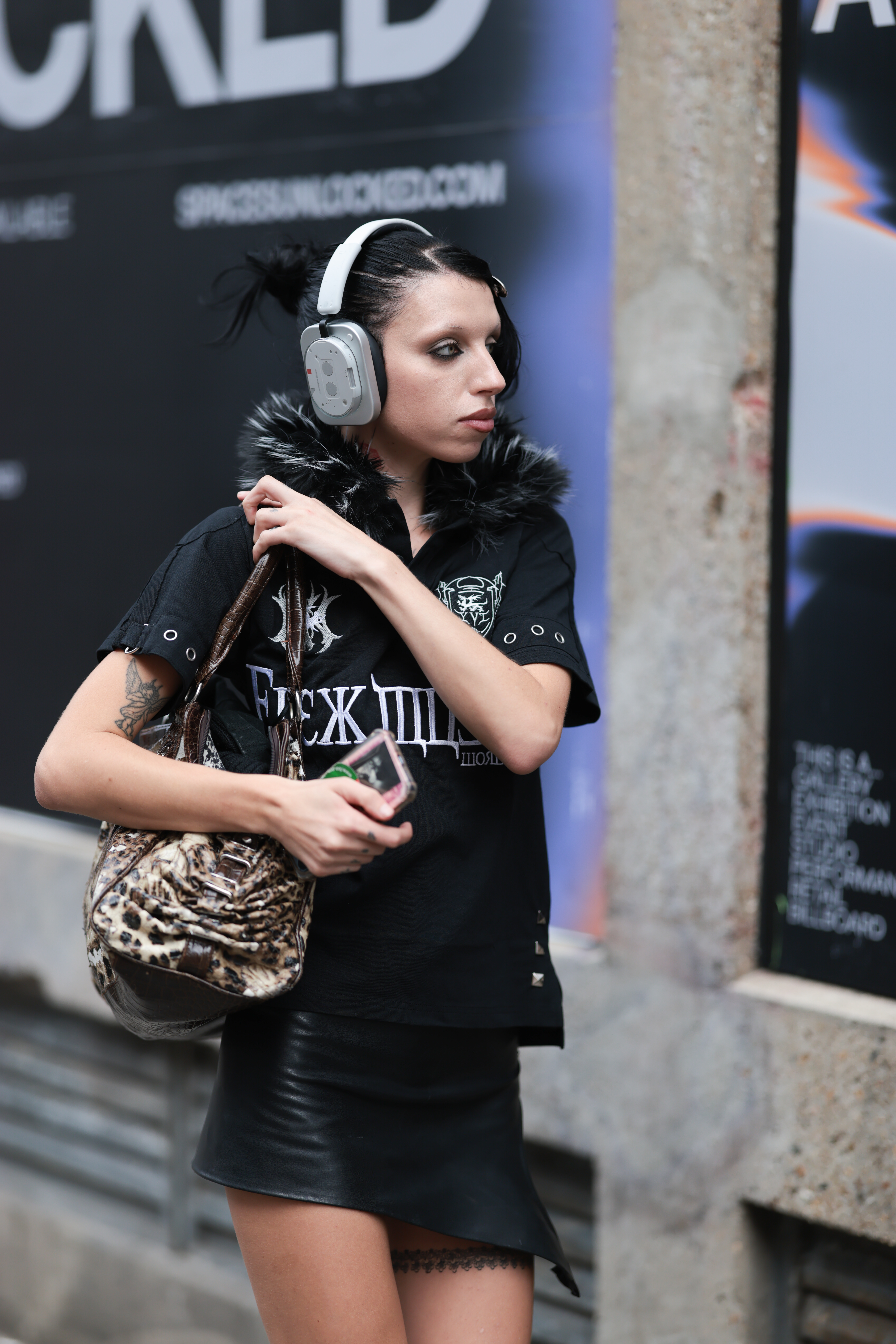 a woman wearing a black t-shirt, black shorts carrying a leopard print bag, white headphones with bleached eyebrows and black eyeliner
