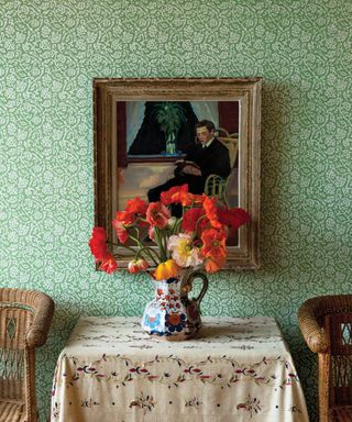 Traditional hallway with green, floral wallpaper from Soane Britain, table with cream tablecloth and floral detailing, antique jug with red flowers, rattan chairs either side of table
