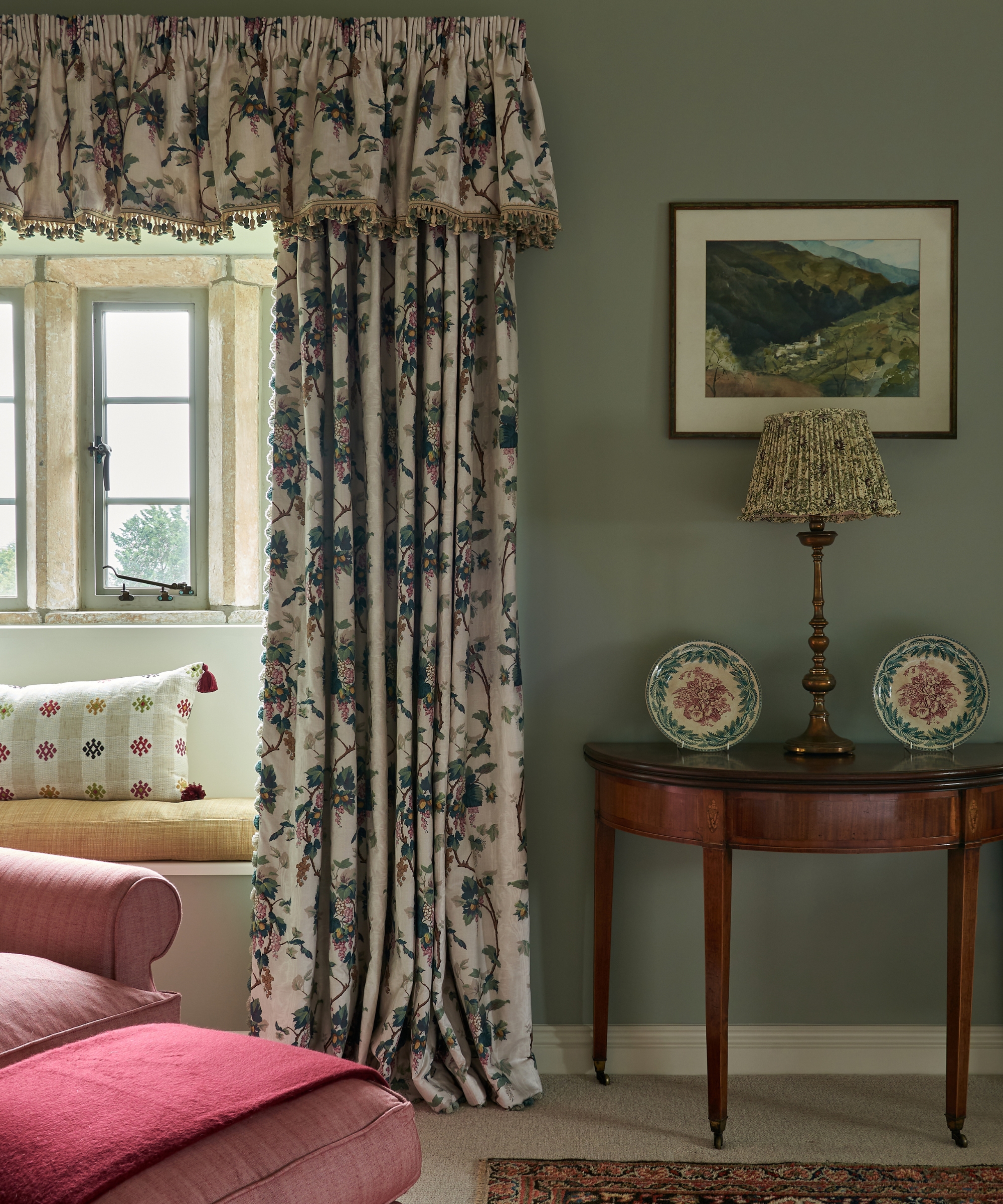 A sitting room with sage-green walls, floral curtains, a patterned lampshade, antique furniture and a pink chaise longue.