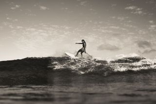 The black and white atmospheric shot shows Malu Kinimaka surfing in the Hawaiian ocean.
