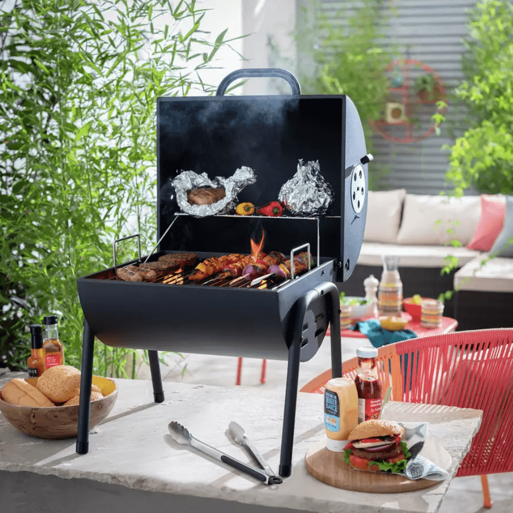 A smaller charcoal BBQ sitting on a counter top with various food cooking on the grill and a burger on the counter