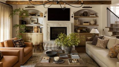 a relaxed farmhouse style family room with arched alcove storage, vintage ceiling beams, neutral couches, fireplace, armchairs and layered foliage