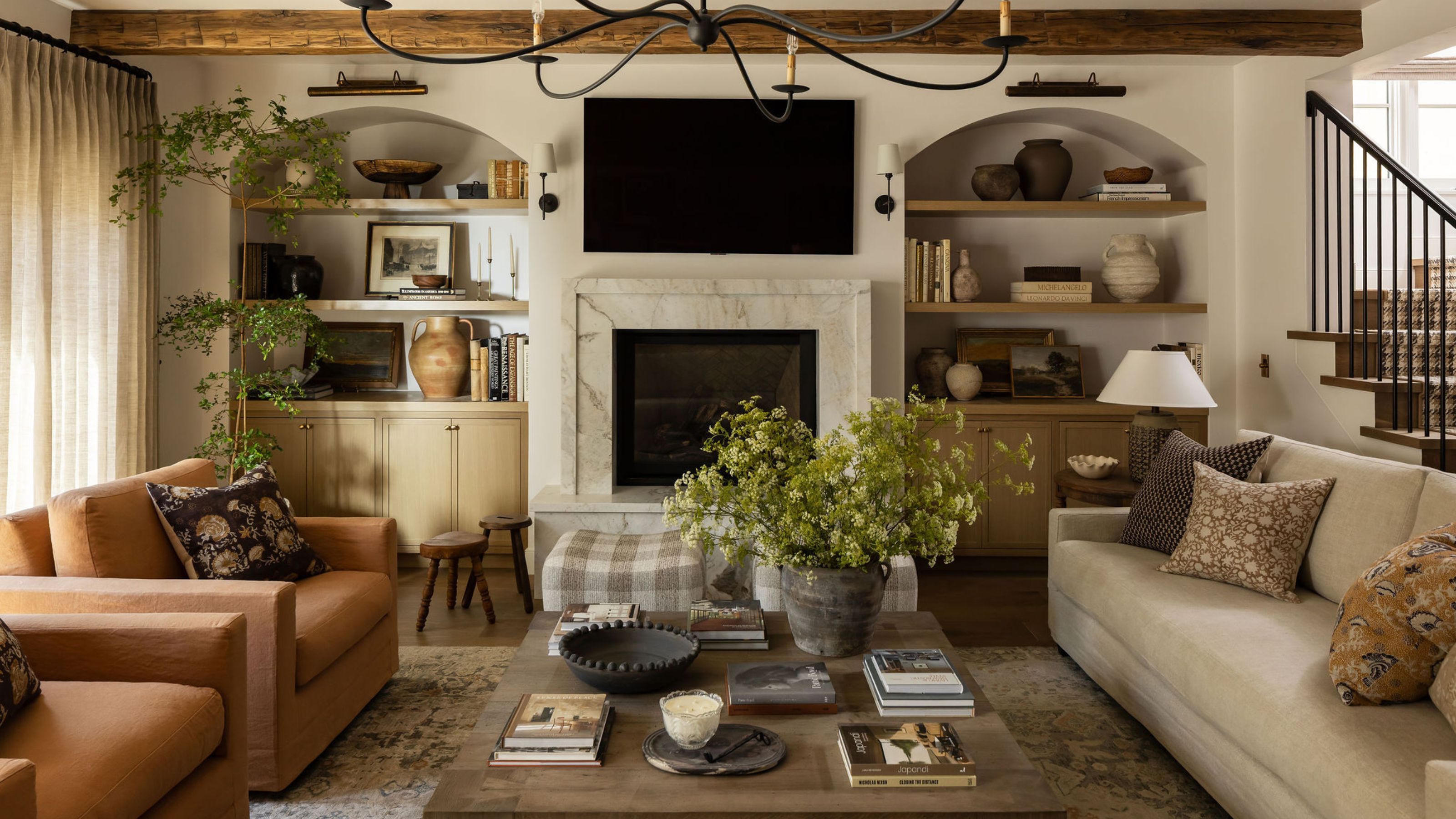 a relaxed farmhouse style family room with arched alcove storage, vintage ceiling beams, neutral couches, fireplace, armchairs and layered foliage