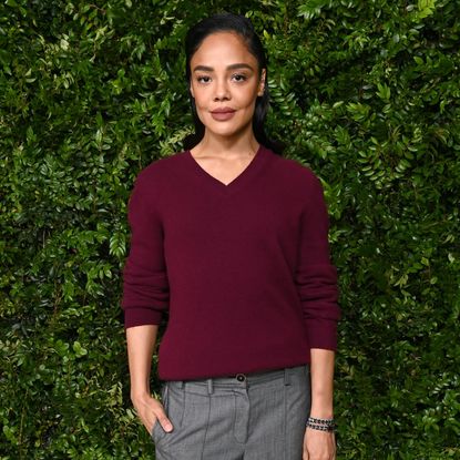 Tessa Thompson standing in front of a plant-decorated backdrop. She is wearing a burgundy sweater and smiling at the camera.