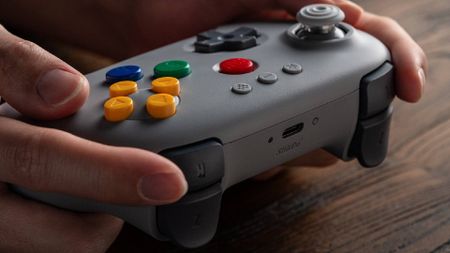 Hands holding 8Bitdo N64 controller in gray resting on woodgrain desk.