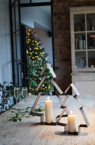 A pair of metal tree shaped candle holders with pillar candles on a table and Christmas tree in background