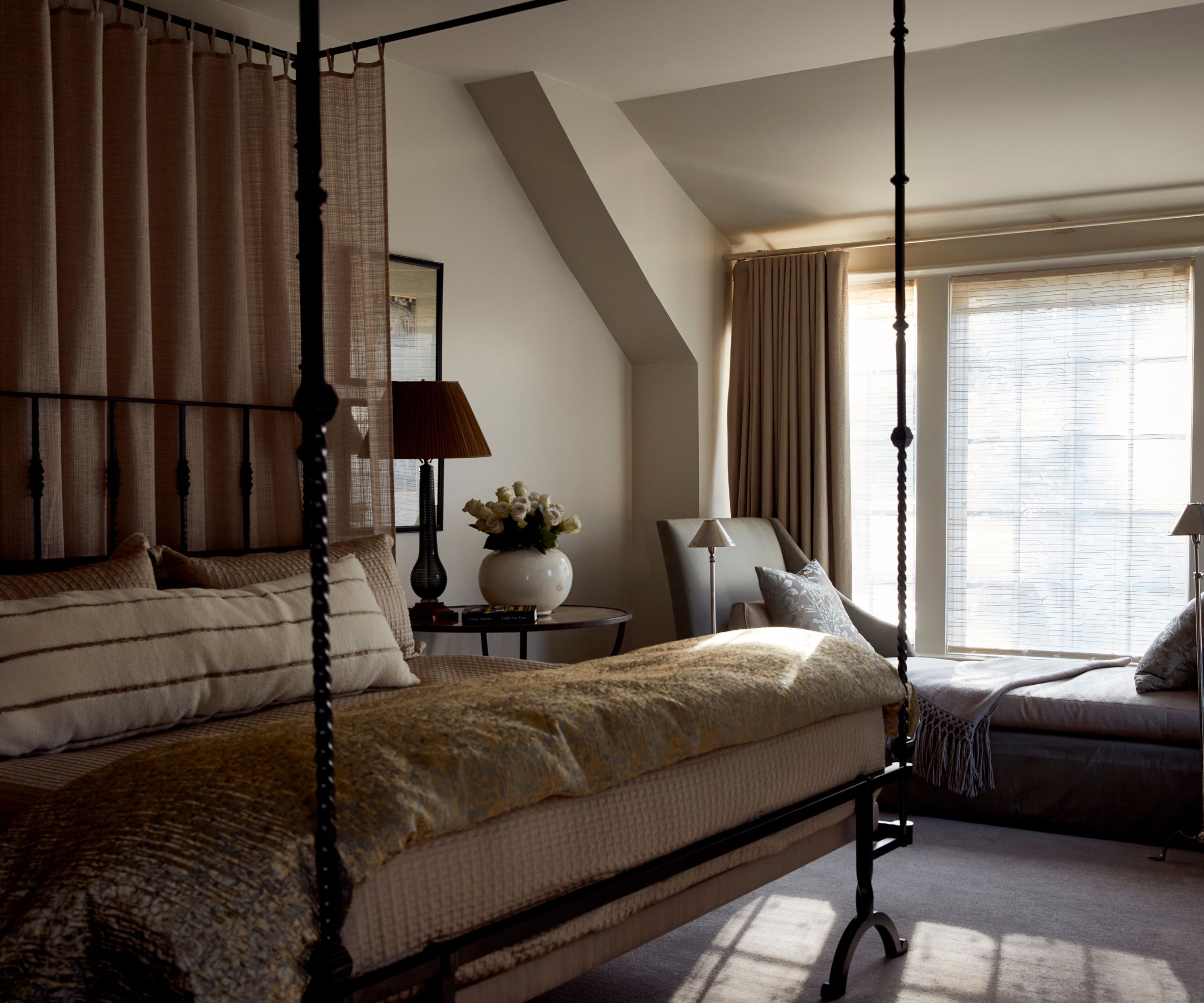 A cozy neutral bedroom with warm white walls, a large window with a bench seat and beige curtains, a carpeted floor, a four-poster canopy bed with layered neutral bedding.