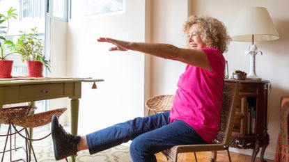 woman wearing a bright pink top and jeans sat on a chair lifting one leg and extending one arm in front. she's in a home setting.