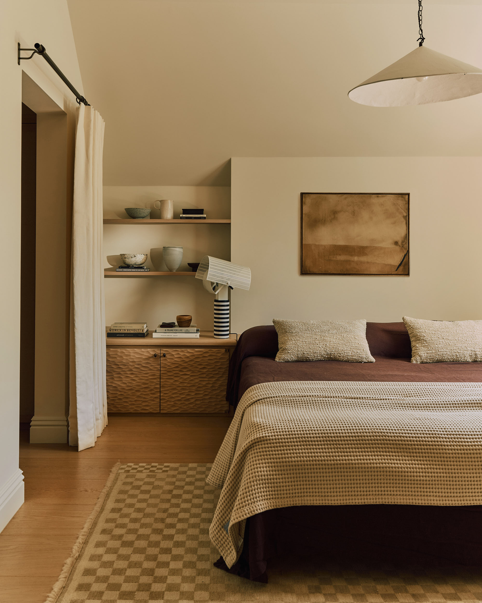 bedroom with brown bed, brown bed sheets, and beige throw, checkered rug, timber floor, curtain doorway, dimpled wood side table with floating shelves and lamp