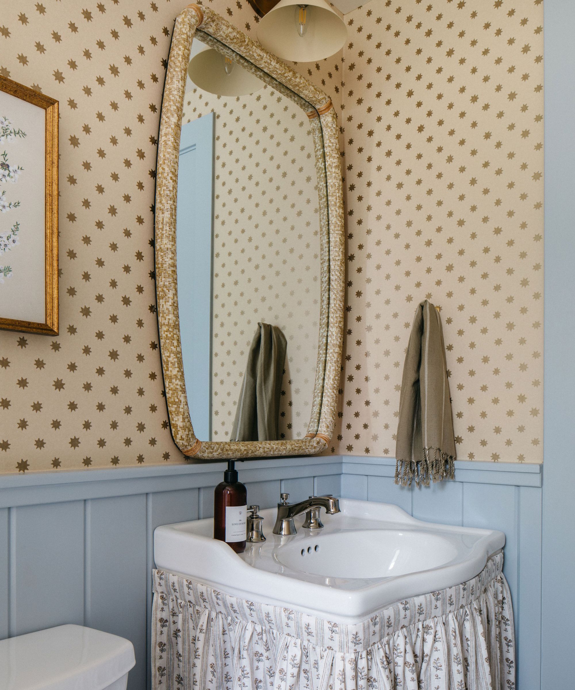 A powder room with powder blue wall paneling, butter yellow patterned wallpaper, a sink with a fabric skirt and a mirror. blue and yellow bathroom