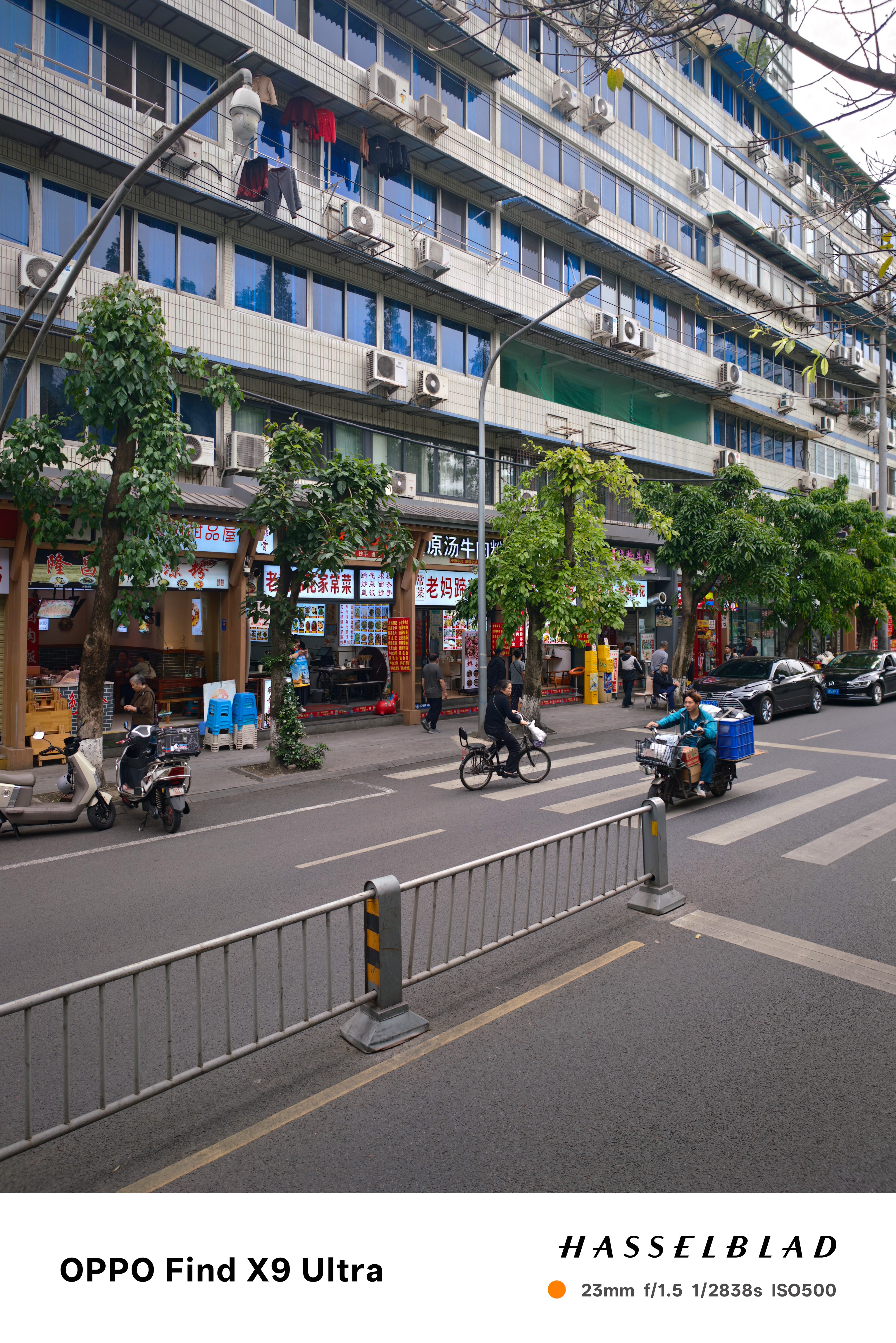 City street lined with shops, trees, scooters, and apartment buildings