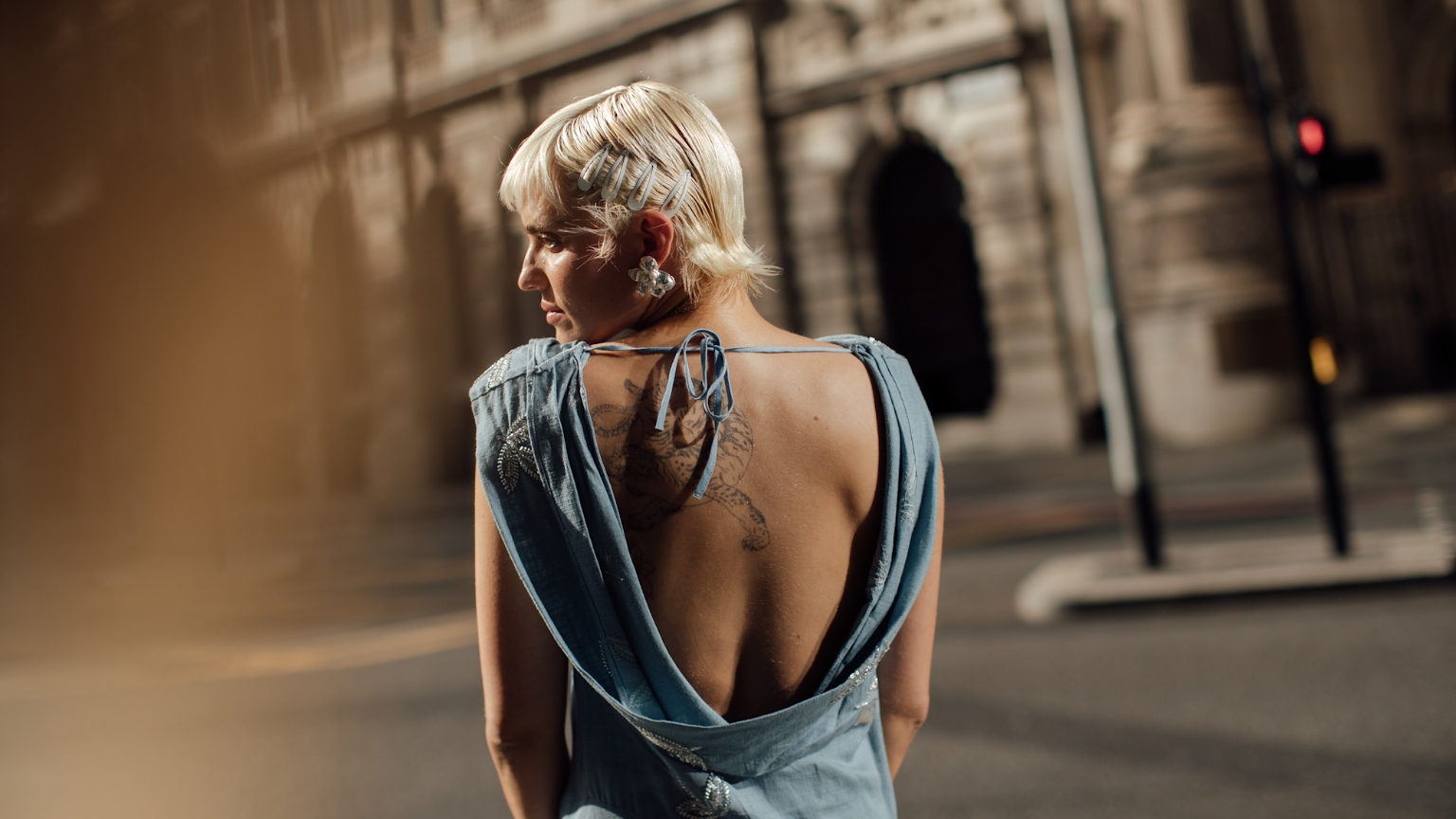 a white woman with a left shoulder tattoo wearing a baby blue backless dress with short platinum blonde hair looking over her shoulder