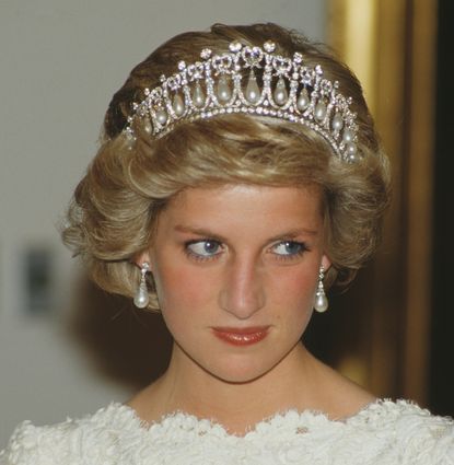 A headshot of Princess Diana wearing a tiara and pearl earrings with a white lace dress