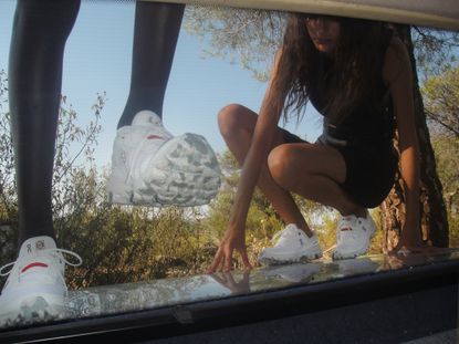 Man and woman in On Running white Loewe sneakers climbing on car in nature