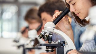 Close up of young woman looking through microscope in science laboratory with other students in the background.