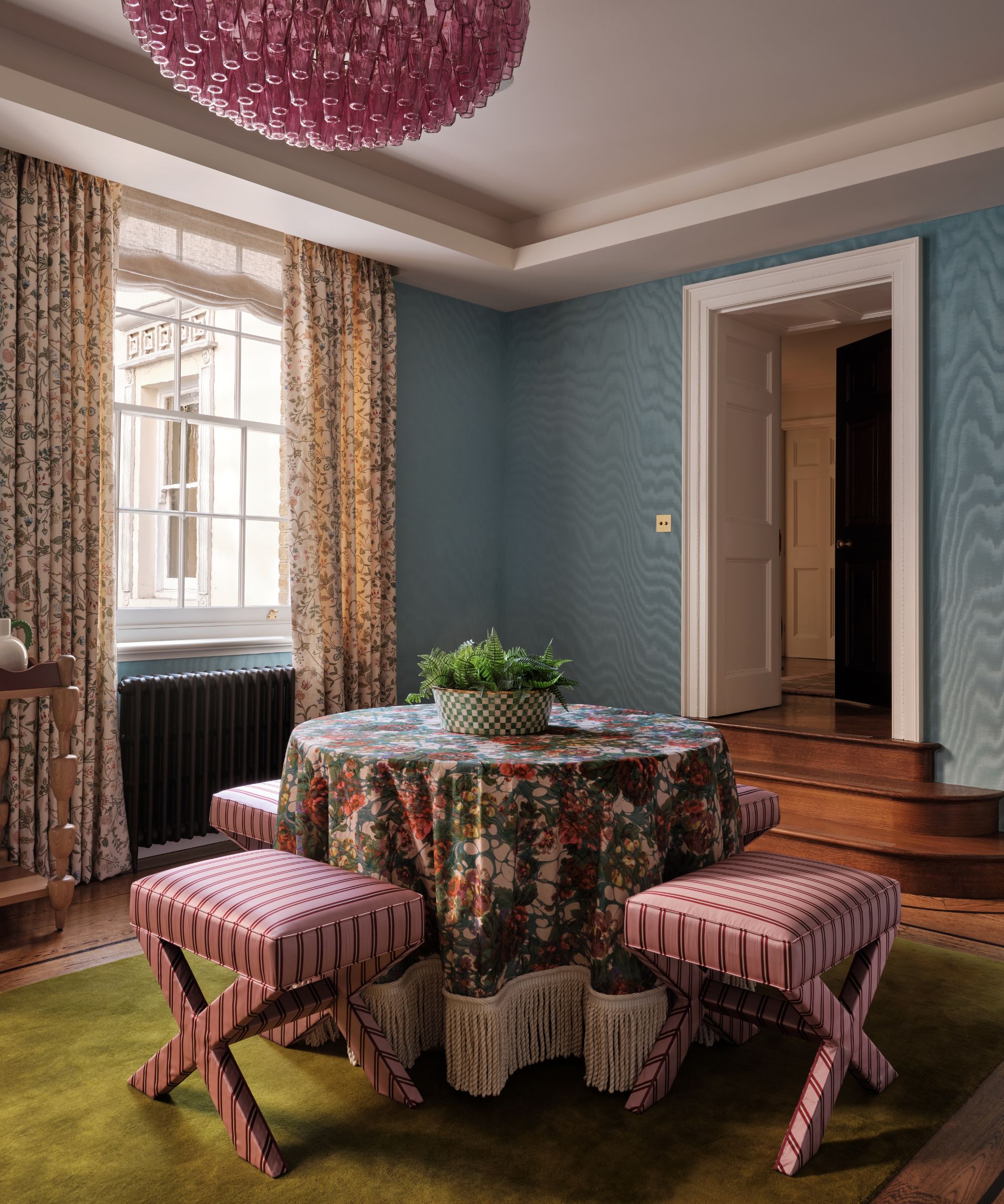 a blue moire wallpapered home library with wooden floor, a green plush rug and a round table with fringed floral tablecloth and striped stools