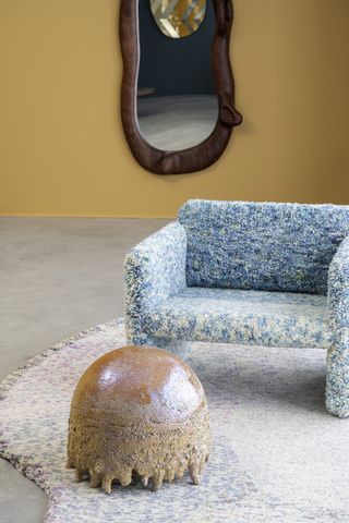 A series of whimsical furniture pieces includes a wooden mirror, a tufted armchair in blue and soft yellow tones, a rug designed in a similar fashion, and a jellyfish-like coffee table.