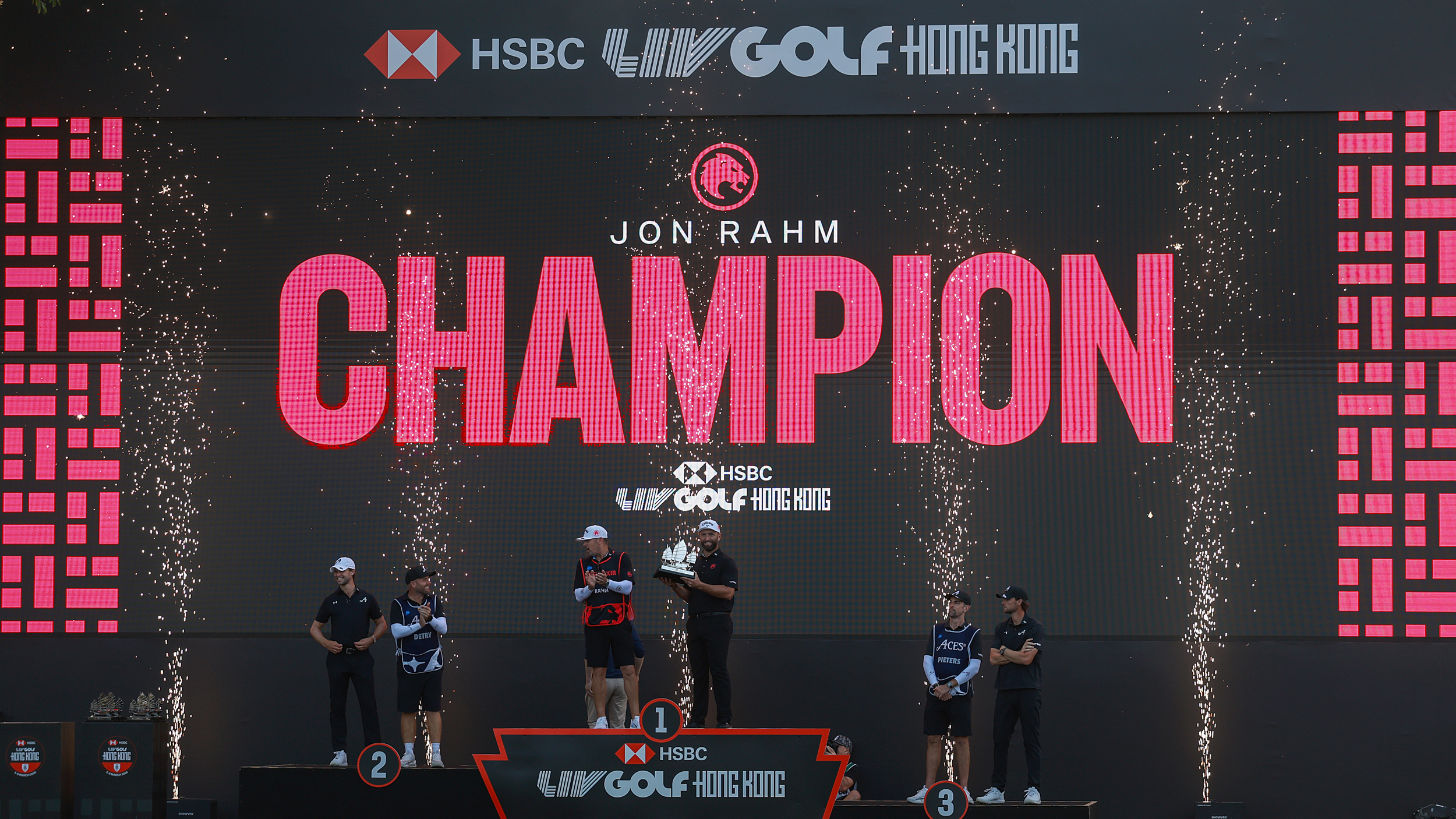 A wide shot of the podium at LIV Golf Hong Kong 2026 with Jon Rahm holding the trophy and Thomas Pieters in third and Thomas Detry in second