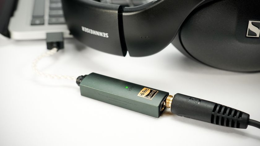 A green iFi Go Link Max lying on a white surface plugged into a black pair of Sennheiser over-headphones and a laptop.