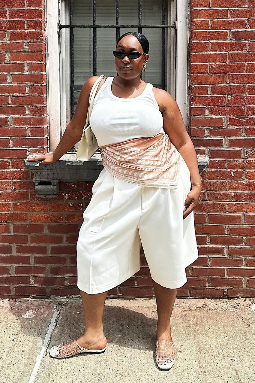 A woman wearing a white tank top, white long shorts, clear jelly shoes, and a printed scarf.