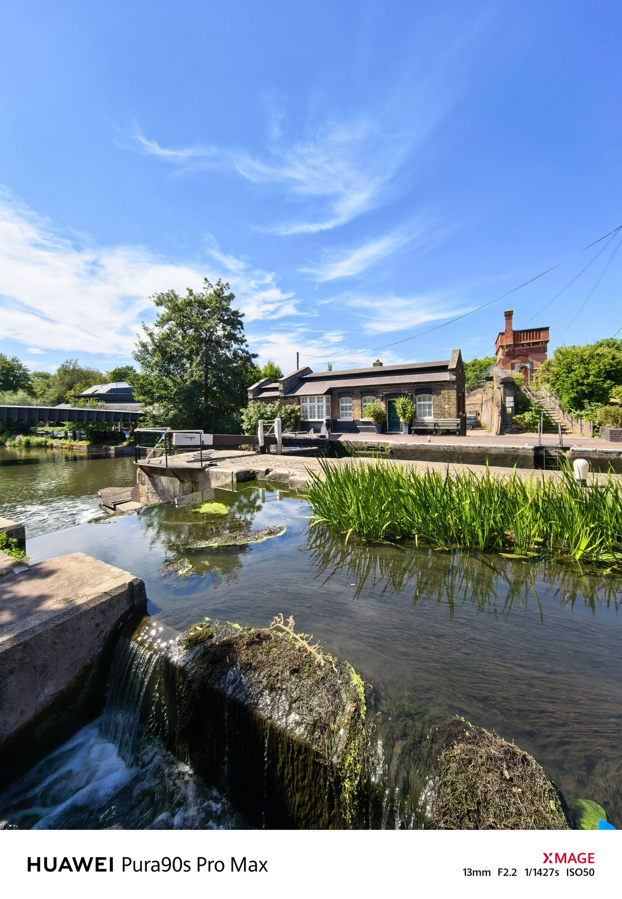 Wide view of a canal lock with flowing water, a lock keeper&rsquo;s cottage and a red-brick building beyond.