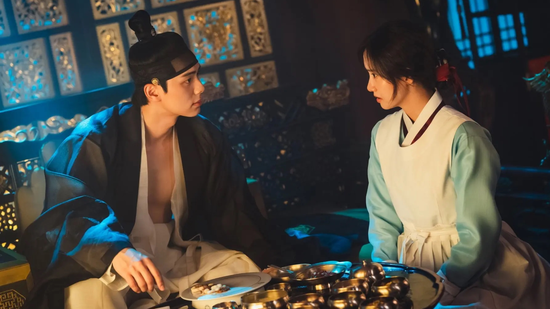 A man (Lee Chae-min) and a woman (Lim Yoon-a) wearing Joseon-era Korean clothing sit in a room in front of a table full of gold, in 'Bon Appetit, Your Majesty.'