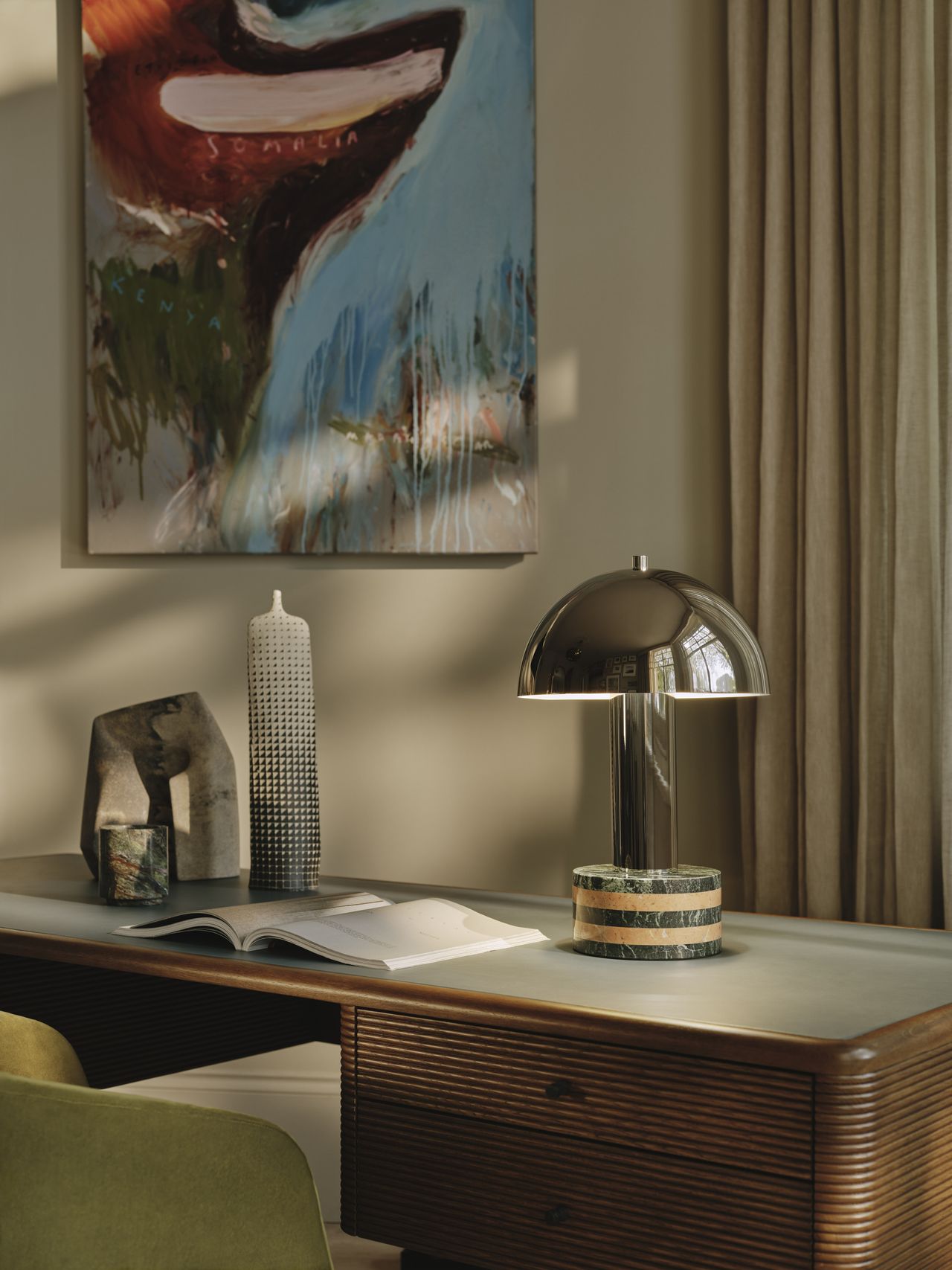Image of a wooden, fluted desk with a light gray top. There is an open book on the desk and a chrome mushroom lamp that has a green and tan, striped marble base. There is an abstract painting on the wall and a light green desk chair in view.