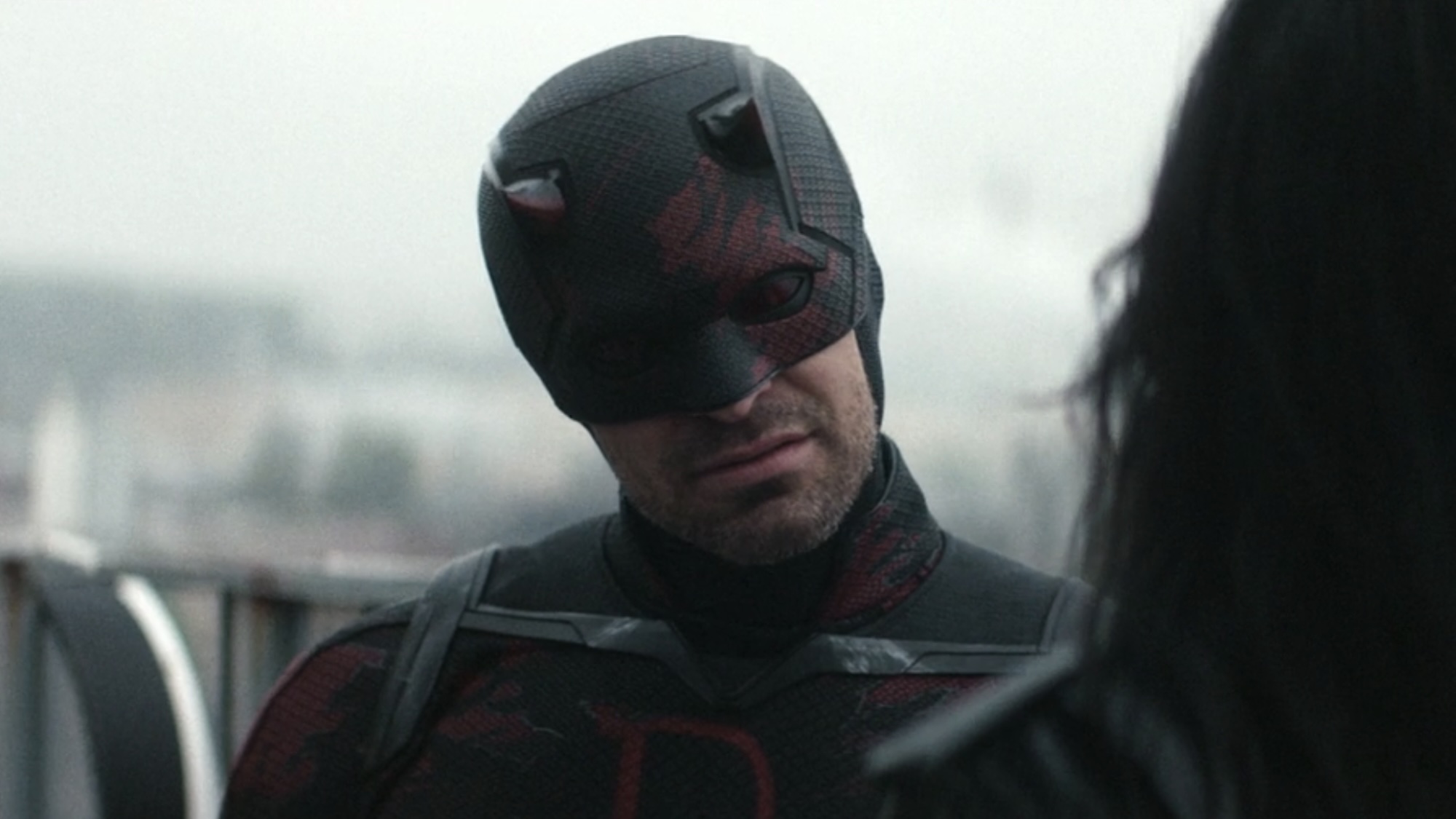 Daredevil in his black and red suit in Season 2 of Born Again, speaking on a rooftop to Jessica Jones during the day. 