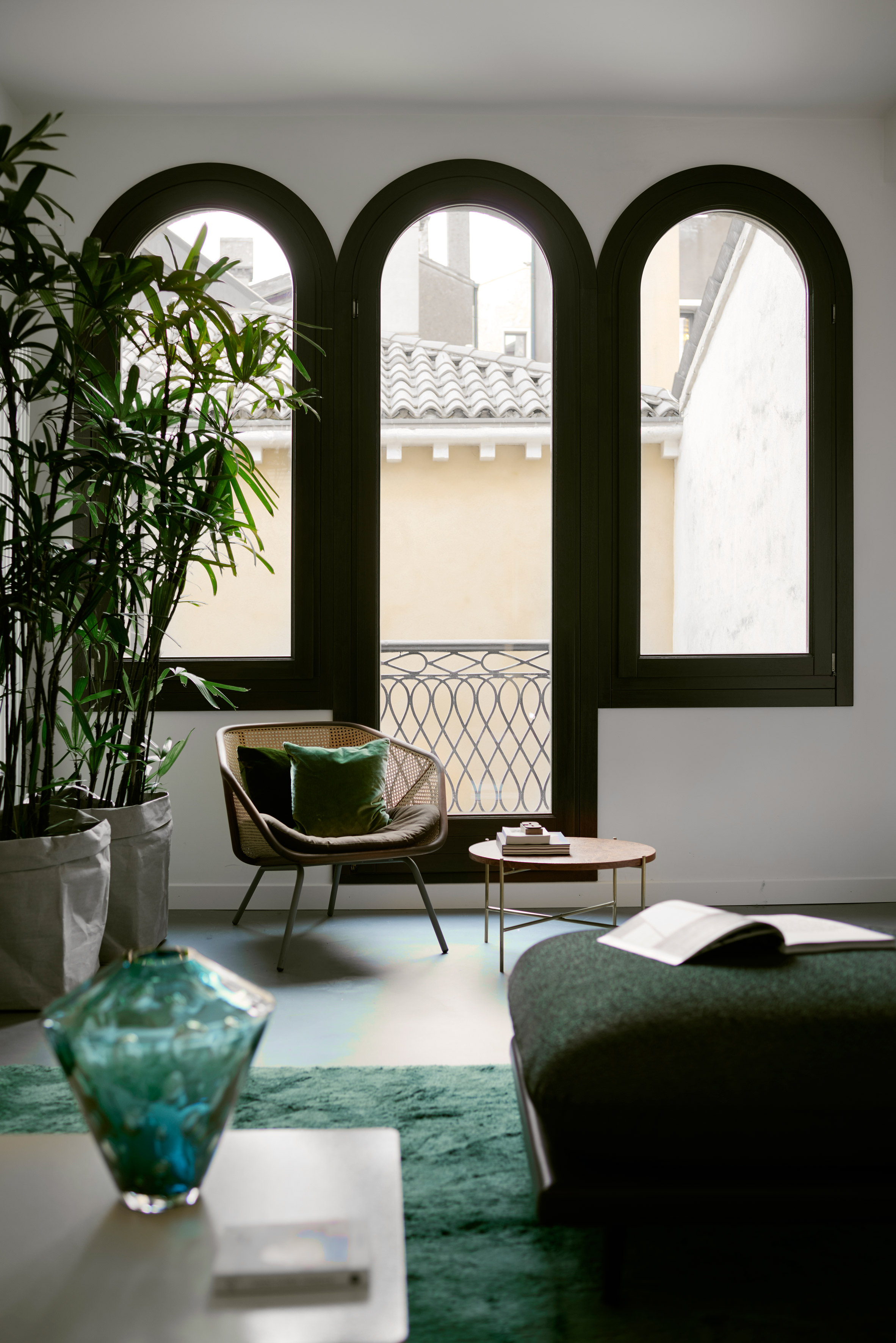 A green-hued apartment showcases a series of furnishings in the same hue, including a fluffy rug, a rectangular, pouf-like sofa, matching cushions positioned on a rattan chair, a vase, and plants.
