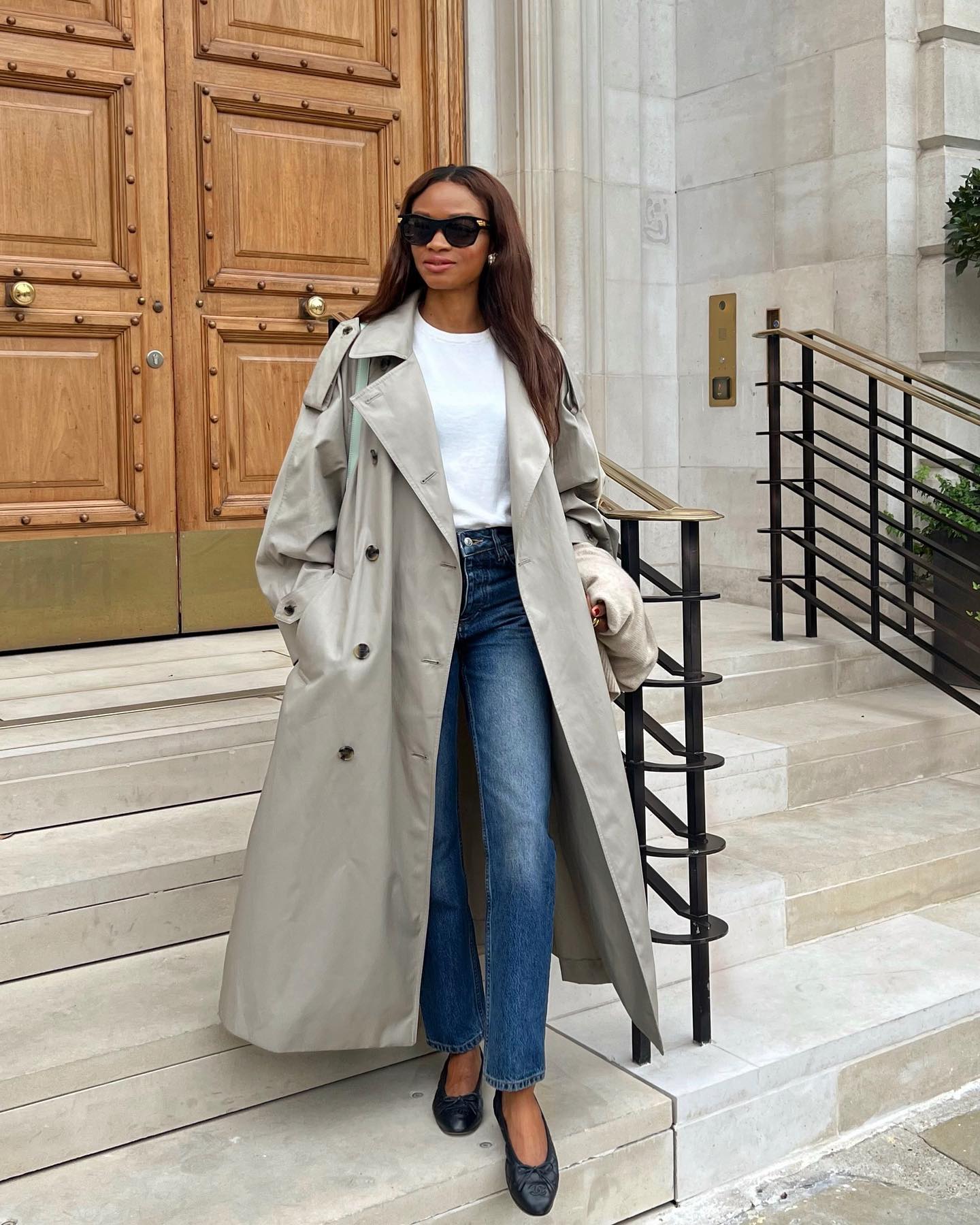stylish French woman poses on steps in Paris wearing black sunglasses, a trench coat, white T-shirt, jeans, and black ballet flats