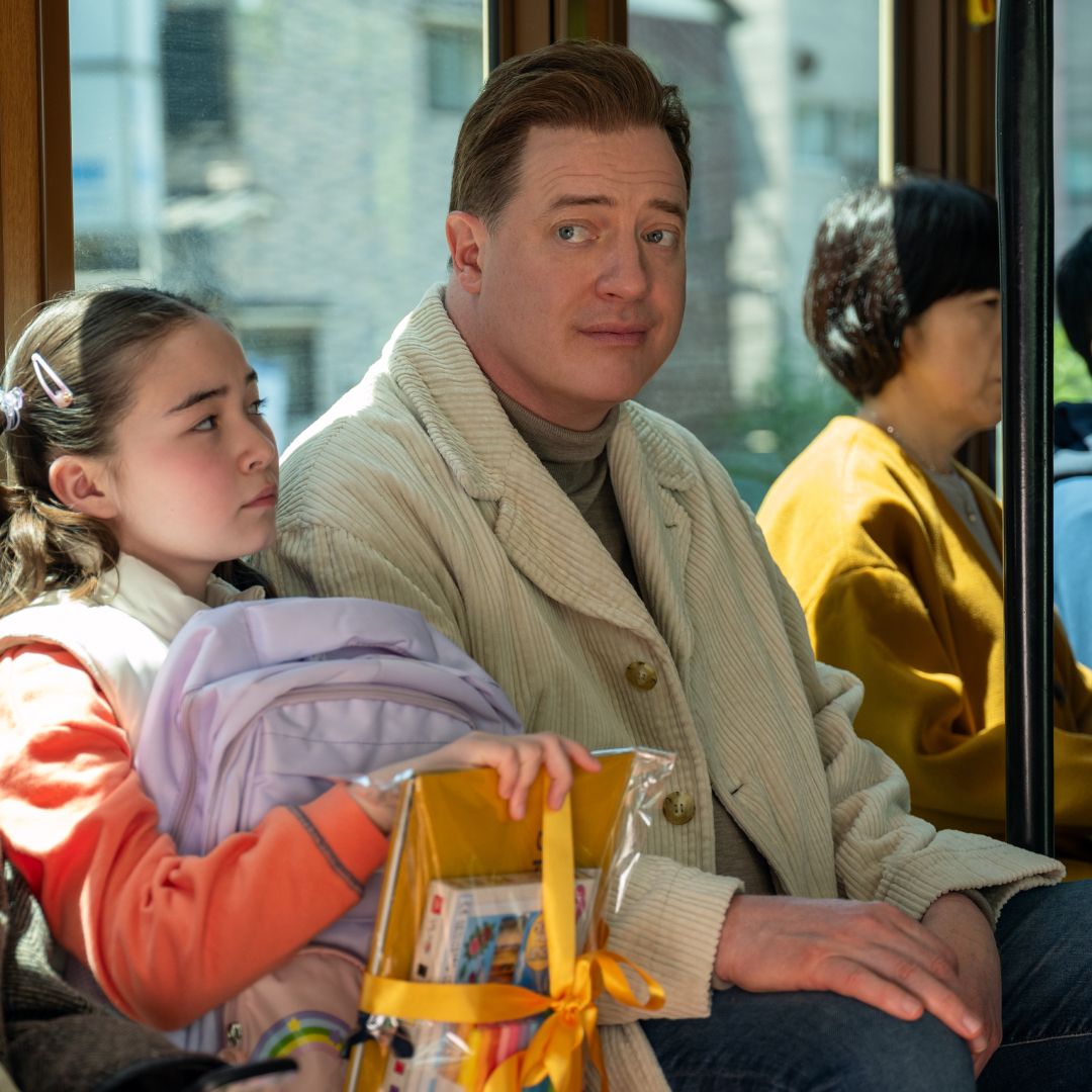 A group of people sitting on a bus. A young girl (Shannon Gorman as Mia) holds a backpack and yellow school supplies, while a man (Brendan Fraser as Phillip) sits next to her, wearing a tan coat. A film still from 'Rental Family.'
