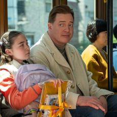 A group of people sitting on a bus. A young girl (Shannon Gorman as Mia) holds a backpack and yellow school supplies, while a man (Brendan Fraser as Phillip) sits next to her, wearing a tan coat. A film still from 'Rental Family.'