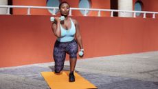 black woman lunging holding two dumbbells in front of red wall on a yellow exercise mat wearing grey leggings turquoise crop top