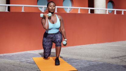 Woman lunging holding two dumbbells in front of red wall on a yellow exercise mat wearing grey leggings turquoise crop top