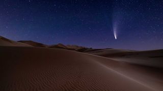 Bright comet in night sky with long white/blue tail. Desert sand dunes are present in the foreground of this image. The sky is also full of stars.